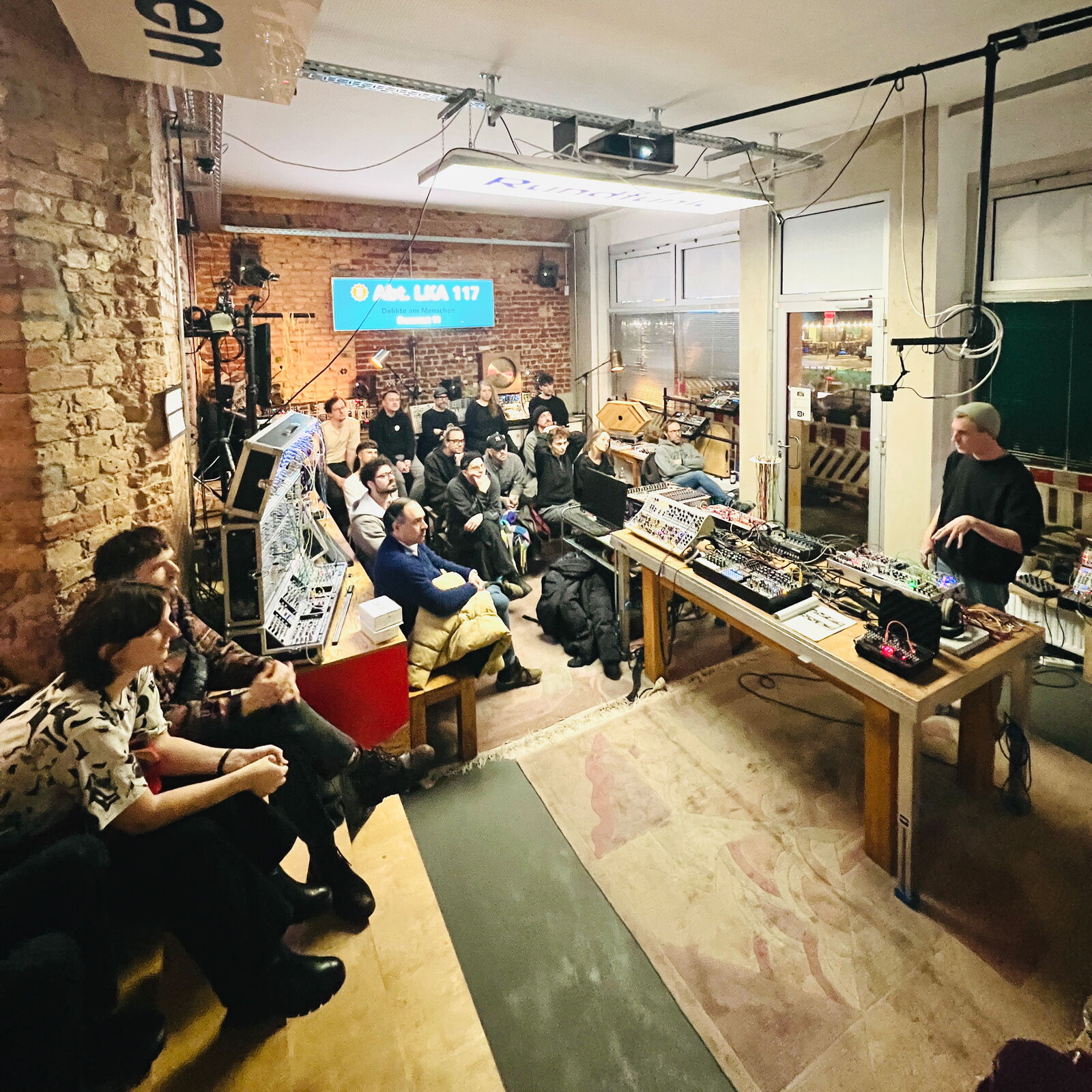 Workshopabend im Berliner SchneidersLaden: Zahlreiche Gäste hören einem Referenten zu, der an einem Tisch voller Modularsynthesizer die Grundlagen erklärt